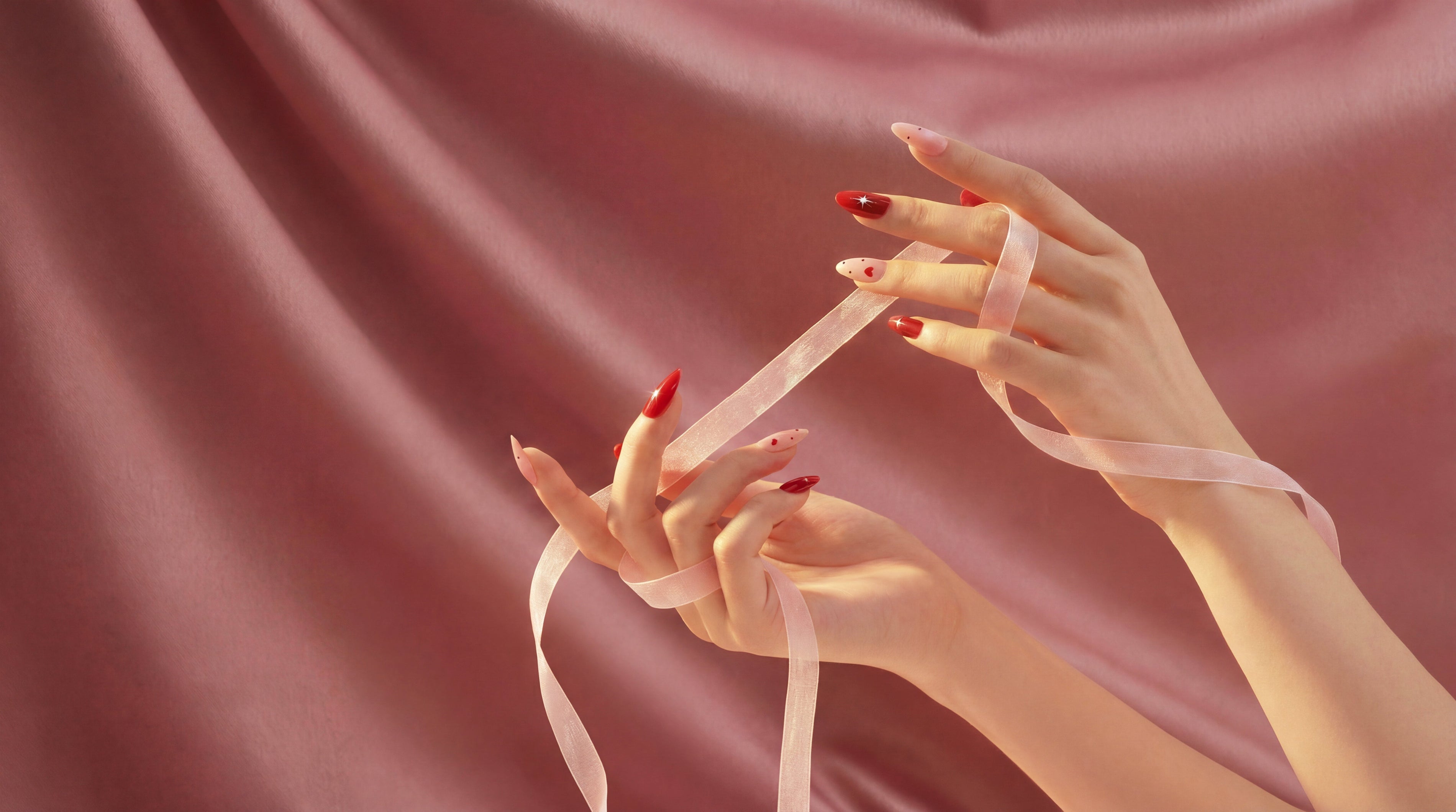 Hand holding pink ribbon with red and nude almond-shaped press-on nails, pink satin background
