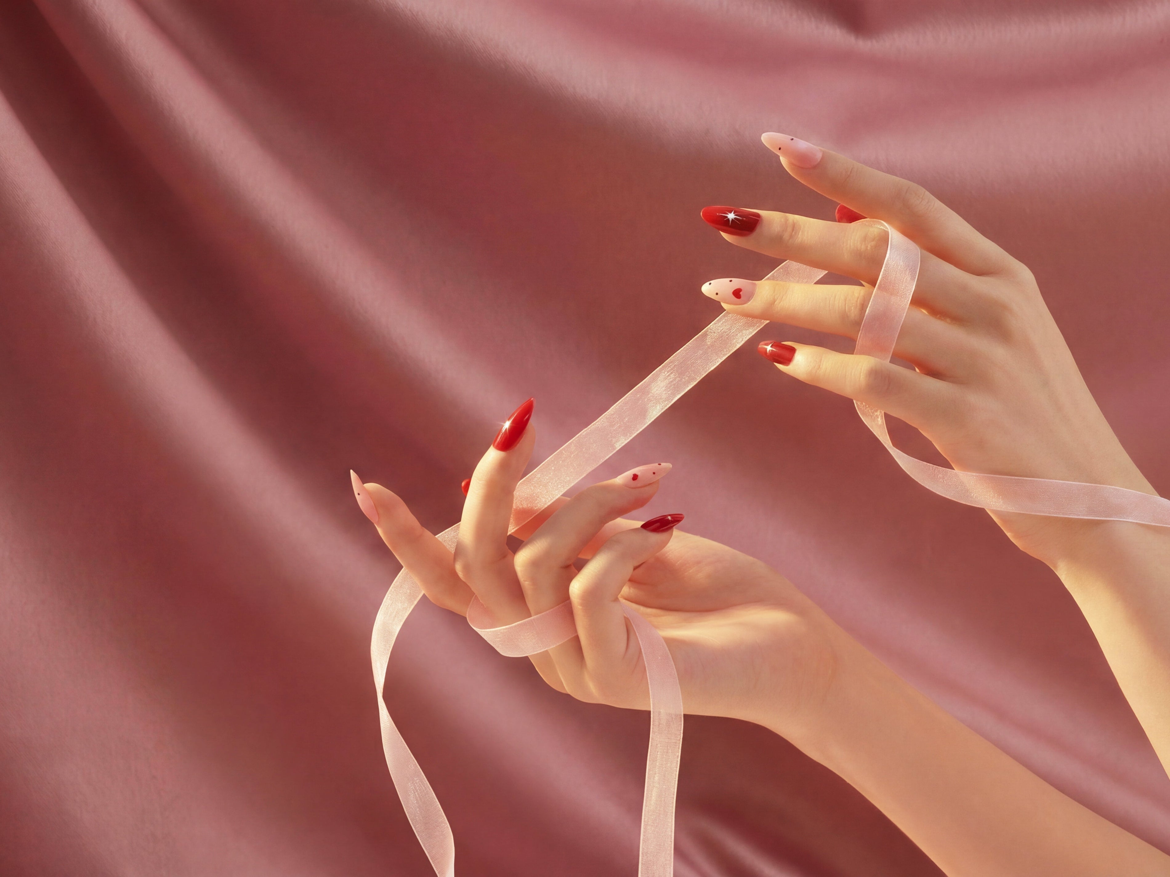 Hand holding pink ribbon with red and nude almond-shaped press-on nails, pink satin background