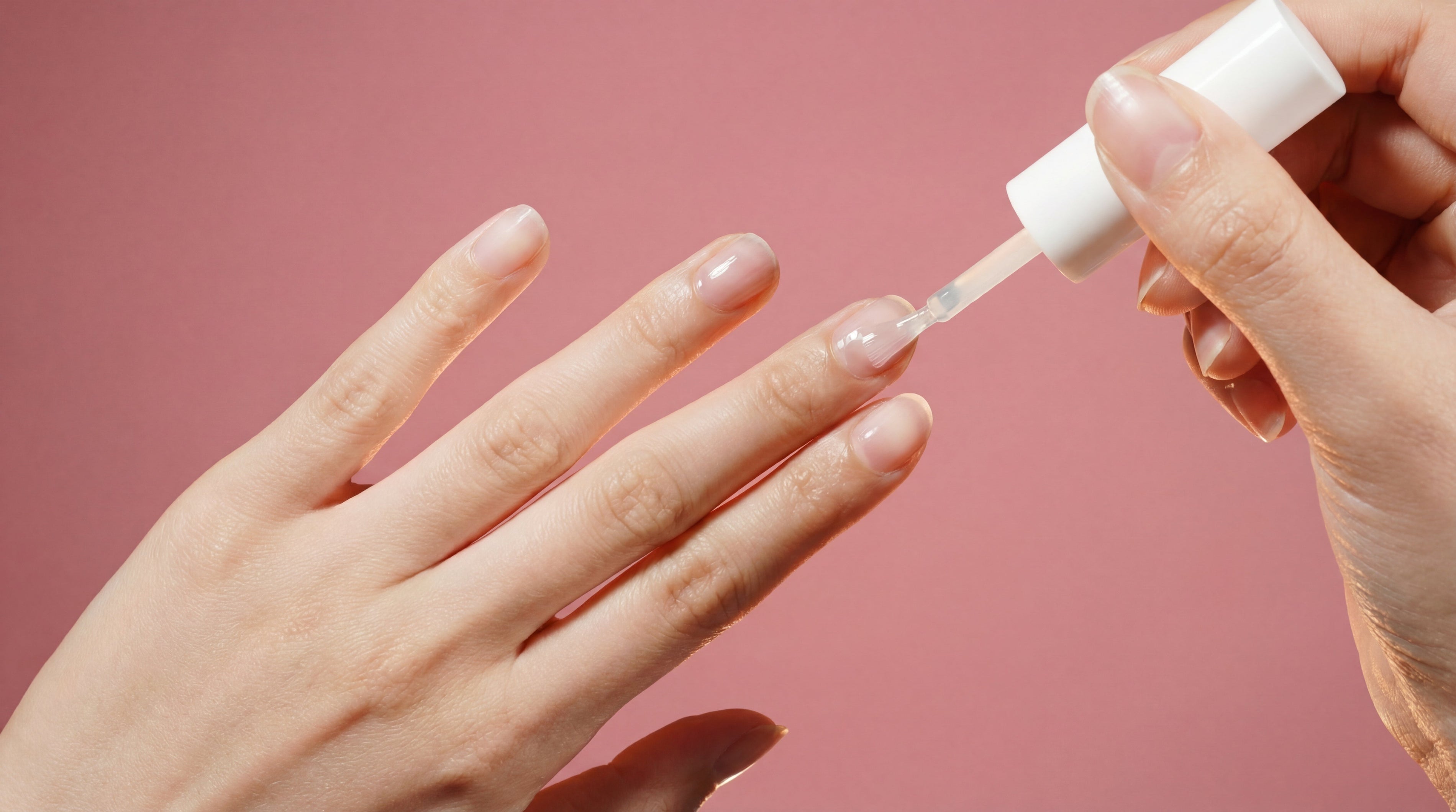Hand applying clear nail polish on natural nails against pink background