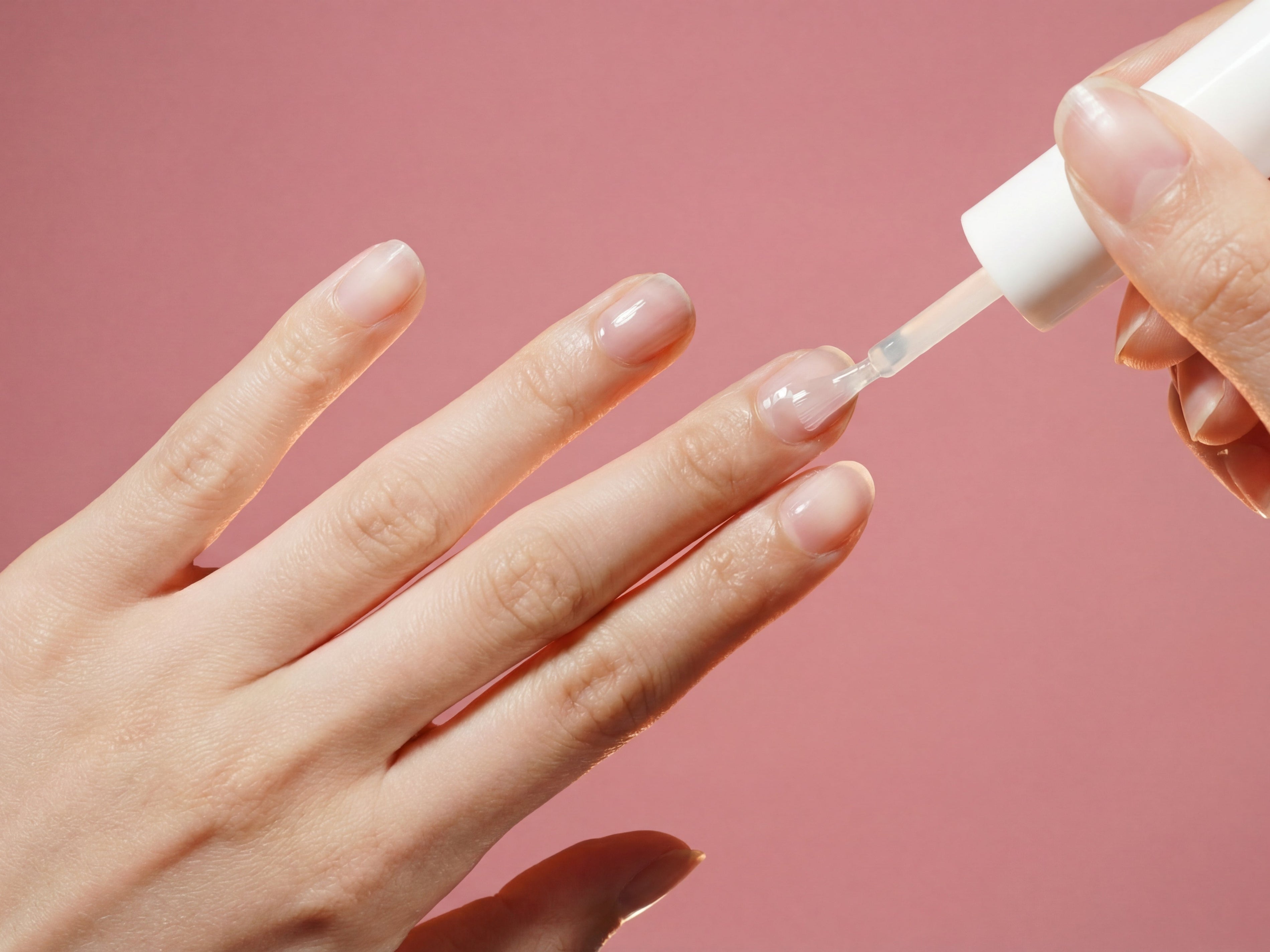 Hand applying clear nail polish on natural nails against pink background