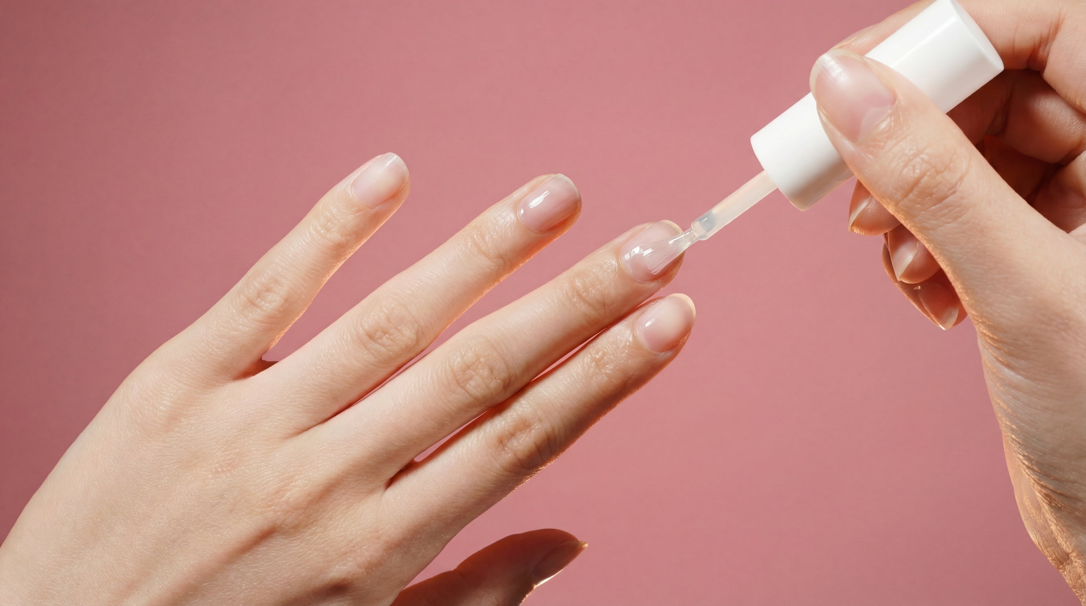 Hand applying clear nail polish on natural nails against pink background