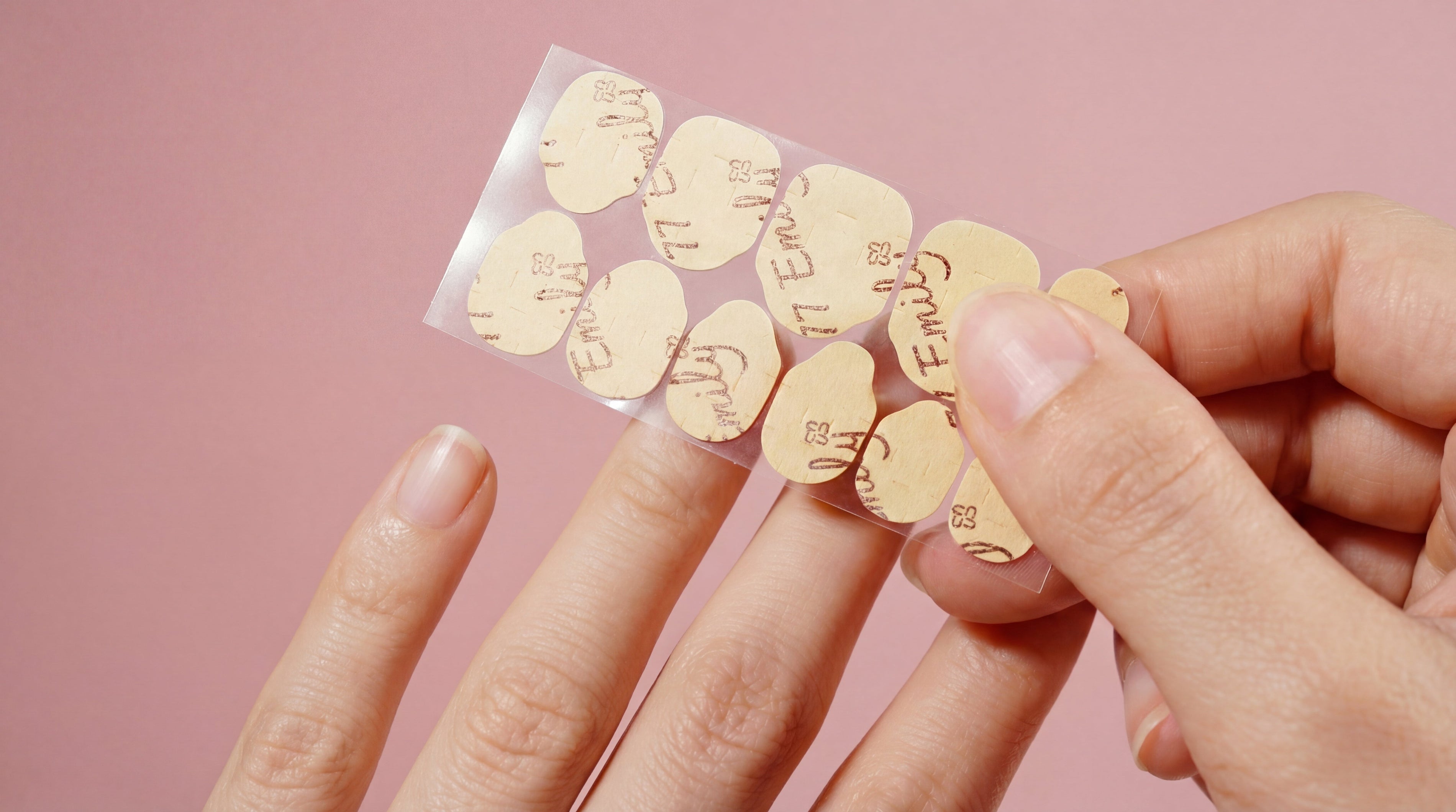 Hand applying adhesive nail tabs for press-on nails with a pink background