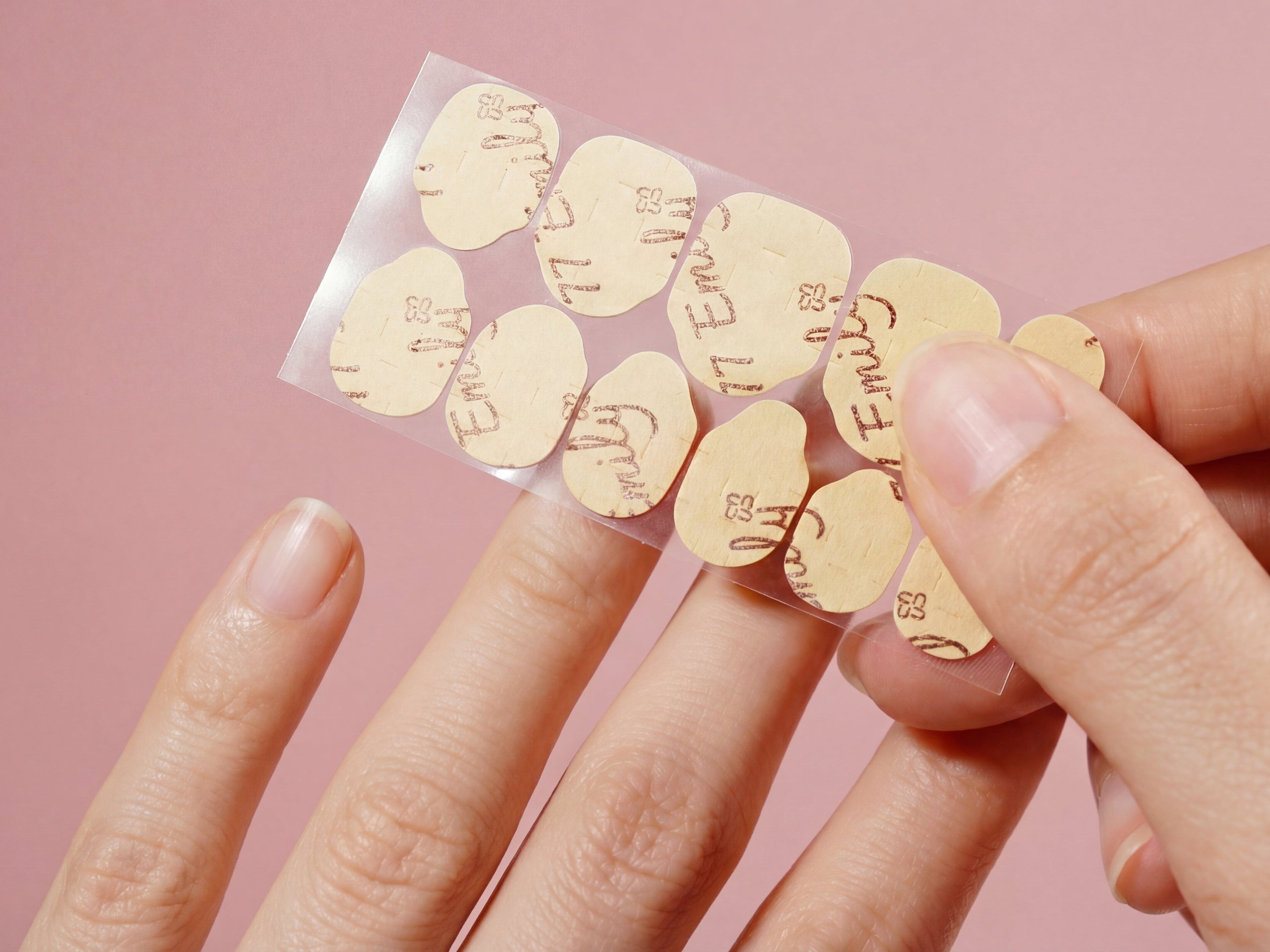 Hand applying adhesive nail tabs for press-on nails with a pink background