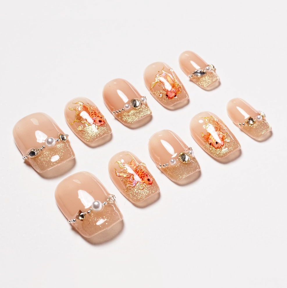 Nude and gold press-on nails with koi fish art, rhinestones, and pearl accents.