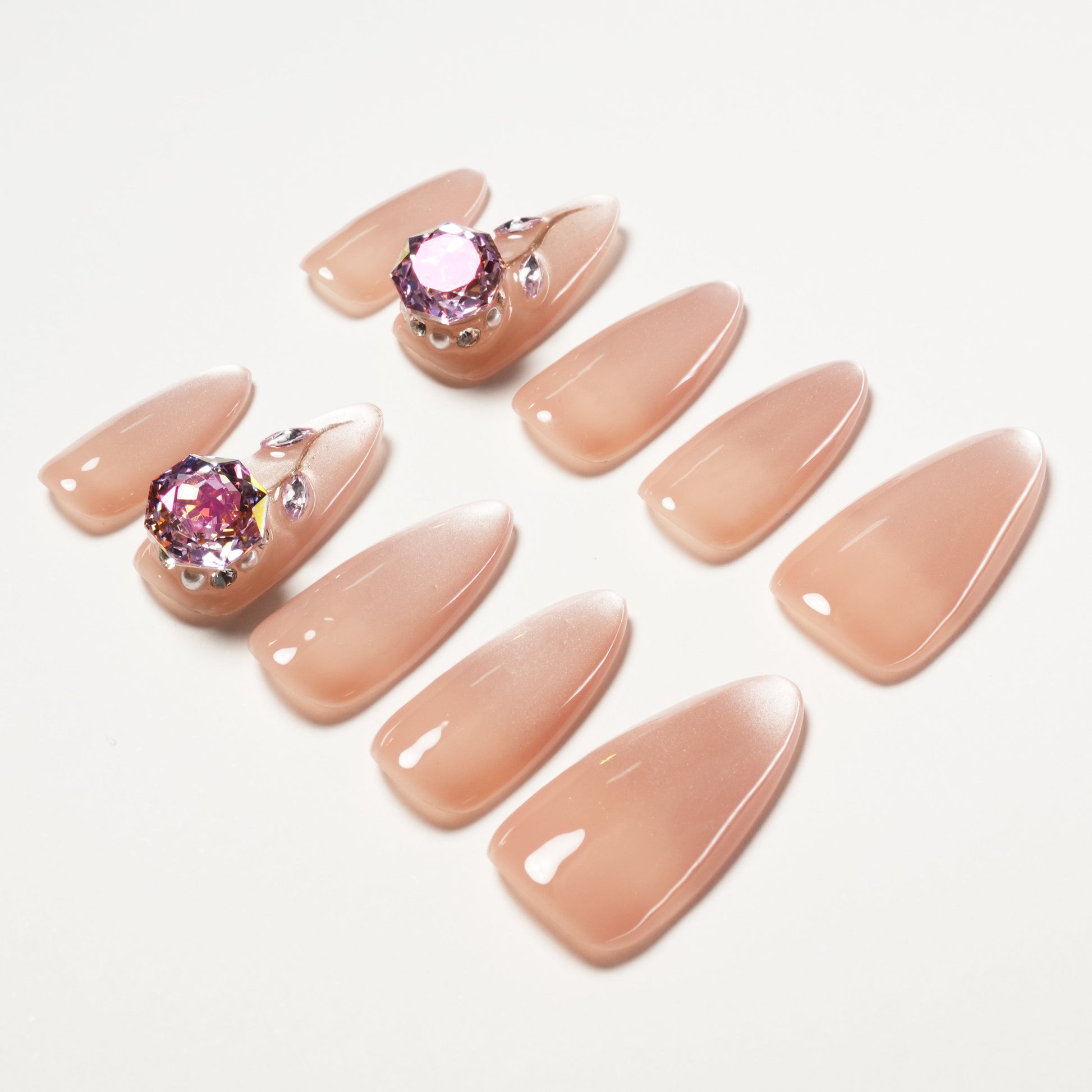 Nude almond press-on nails with pink gem and crystal 3D accents on white background