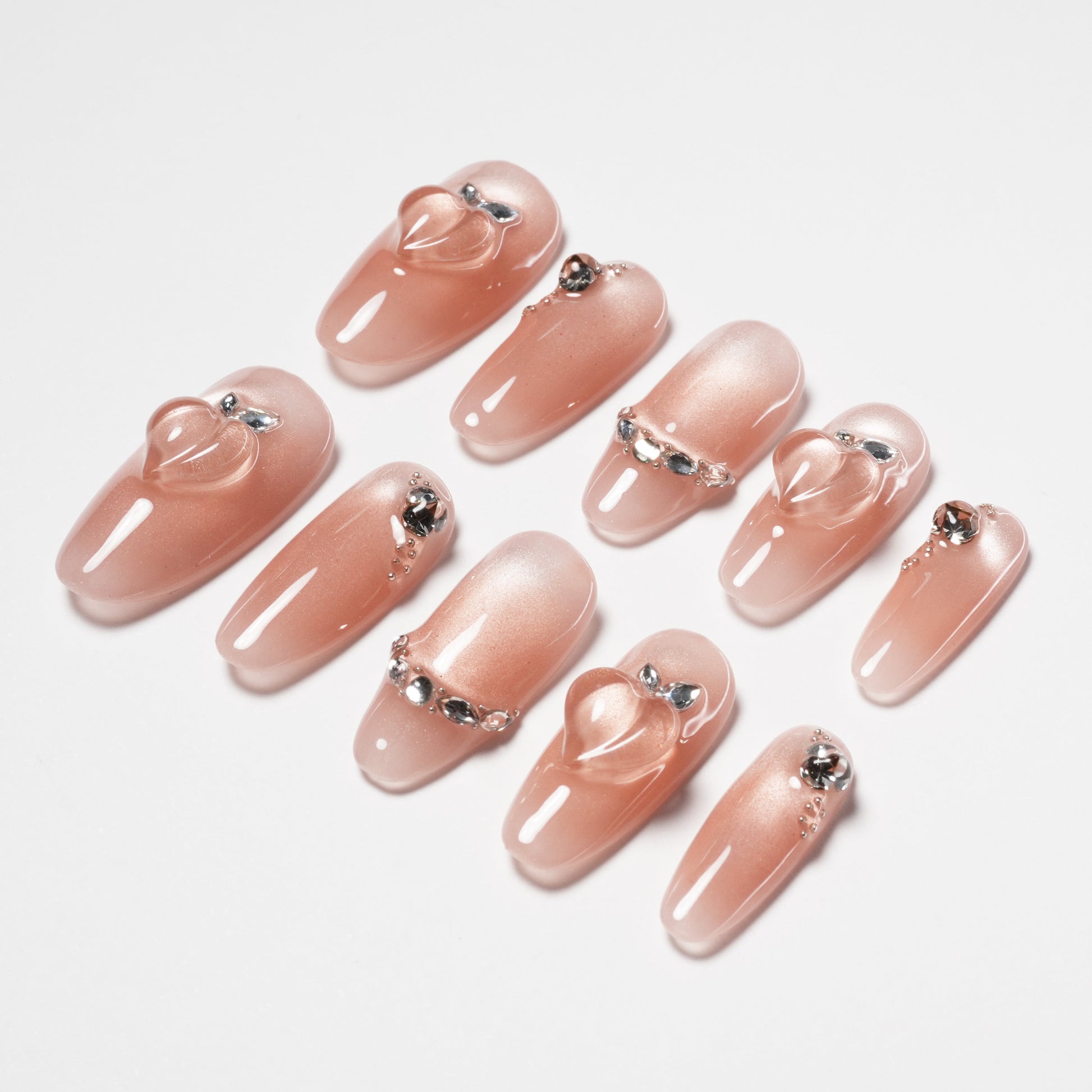 Glossy nude pink press-on nails with heart and rhinestone accents, arranged on white background.