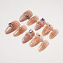 Glossy nude press-on nails with 3D bows, rhinestone gems, and gold glitter accents