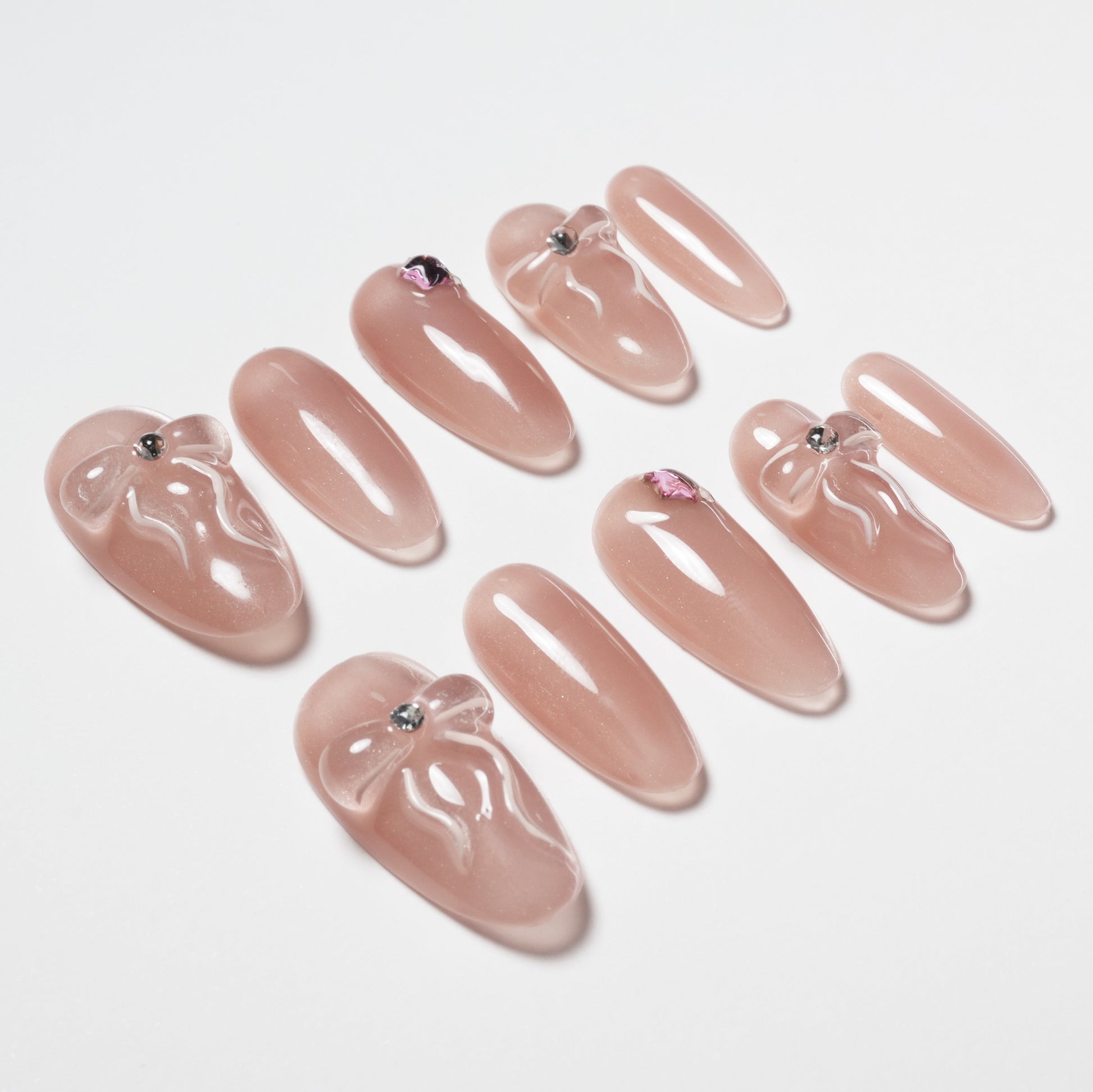 Glossy nude press-on nails with 3D bows and rhinestone accents arranged on white background.