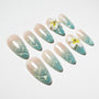 Almond-shaped press-on nails with blue-green ombre, gold shimmer, and 3D floral accents