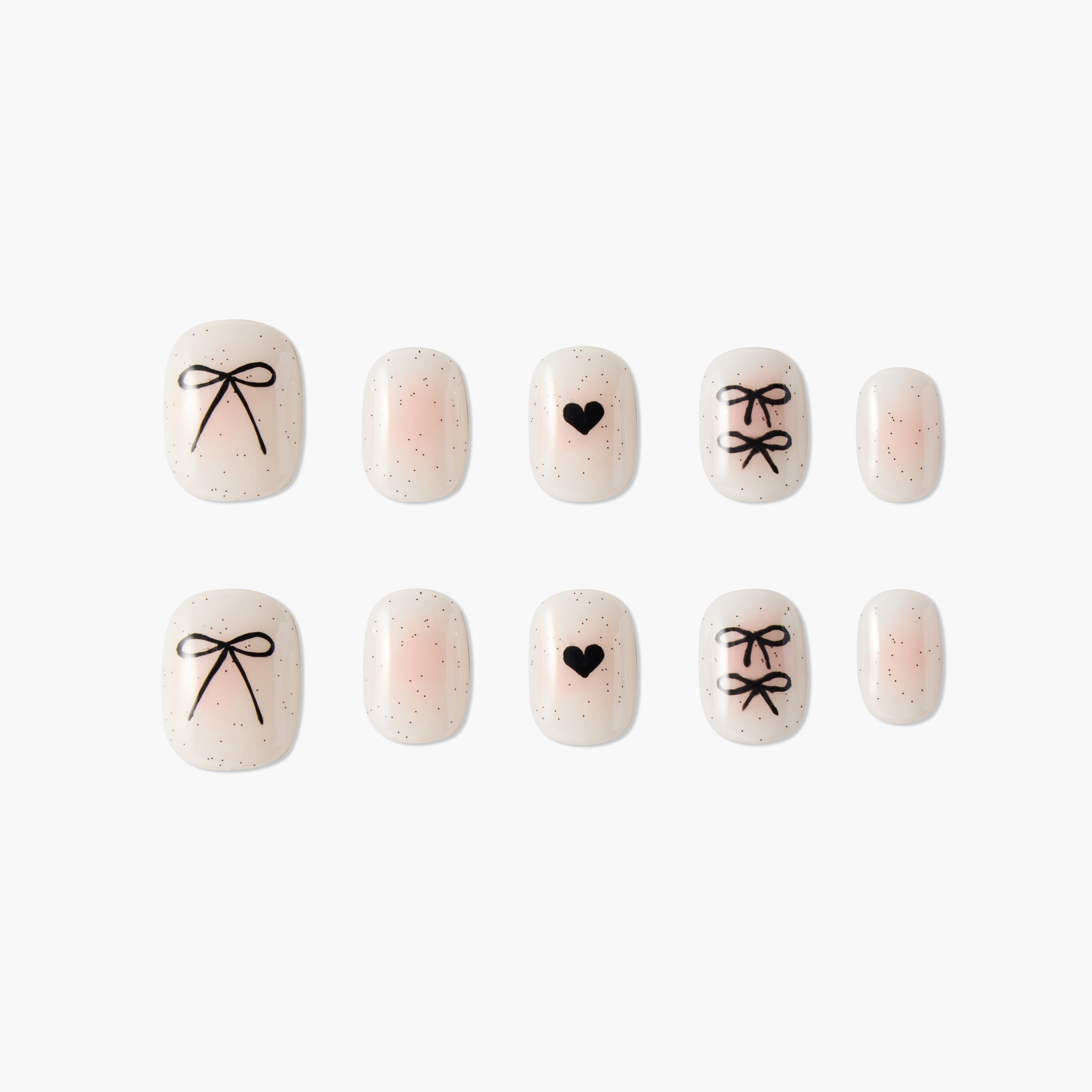 Pink nude press-on nails with black bow and heart designs, glitter accents, on white background.
