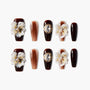 Brown press-on nails with 3D white floral accents, gold gems, and marble design details.
