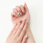 Hand with long almond press-on nails featuring gold, white floral designs, and crystal accents