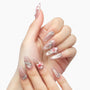Hand with long almond-shaped press-on nails, featuring pink floral 3D designs and rhinestones