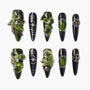 Handmade press-on nails with black base, green 3D floral designs, metallic accents, and rhinestones