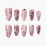 Pink press-on nails with white flower accents, metallic details, and rhinestone embellishments.