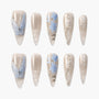 Long almond press-on nails with nude base, blue floral patterns, silver rhinestones, and butterfly accents