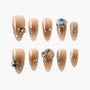 Glossy nude almond press-on nails with 3D rhinestone and chain accents, arranged on white background.