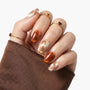 Hand with gold and brown plaid press-on nails, wearing gold rings, brown sleeve visible