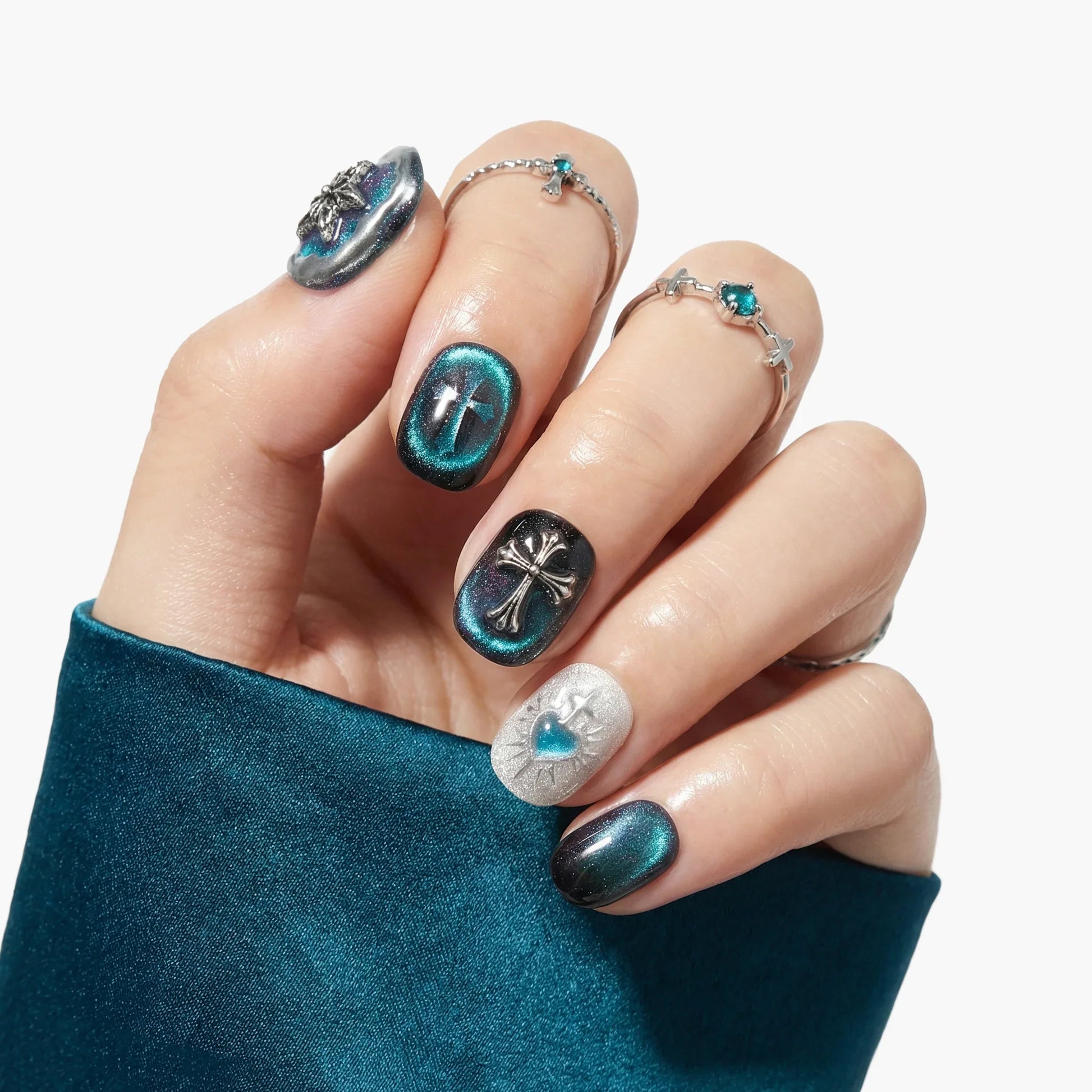 Hand with teal and black metallic press-on nails, cross and heart 3D nail art, and silver rings