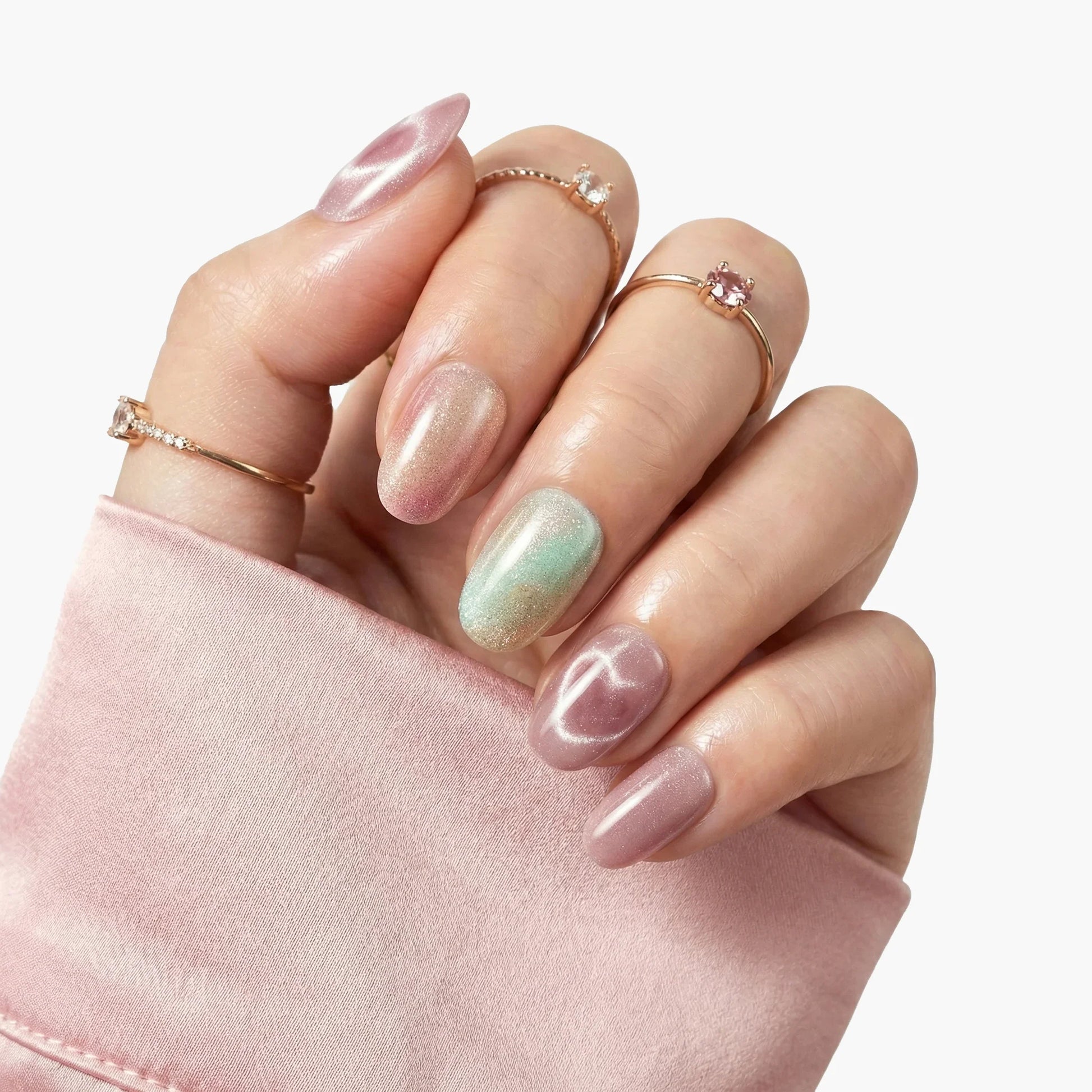 Hand with pastel iridescent press-on nails and gold rings, soft pink sleeve background