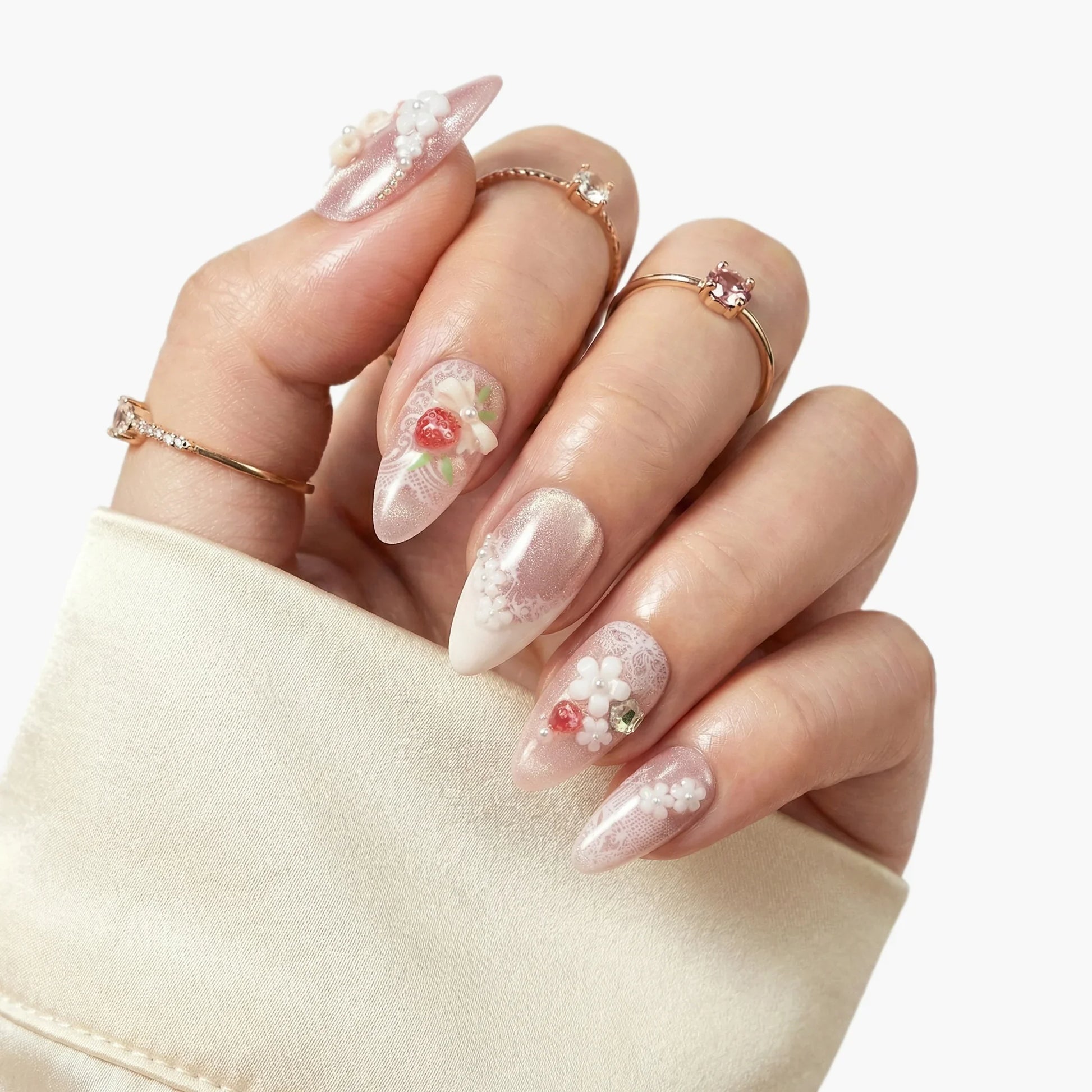 Hand with almond-shaped press-on nails featuring 3D floral designs, pearls, and strawberries.