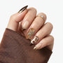 Hand with almond-shaped press-on nails featuring brown, gold, and crystal floral designs.