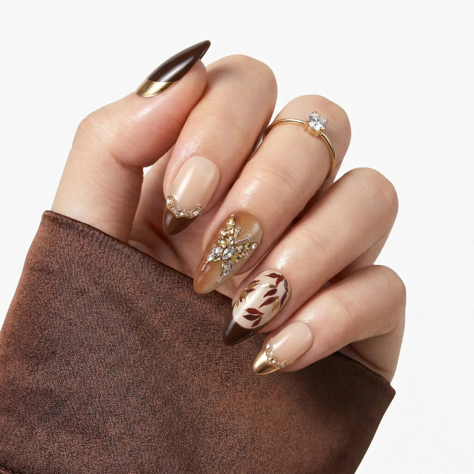 Hand with almond-shaped press-on nails featuring brown, gold, and crystal floral designs.
