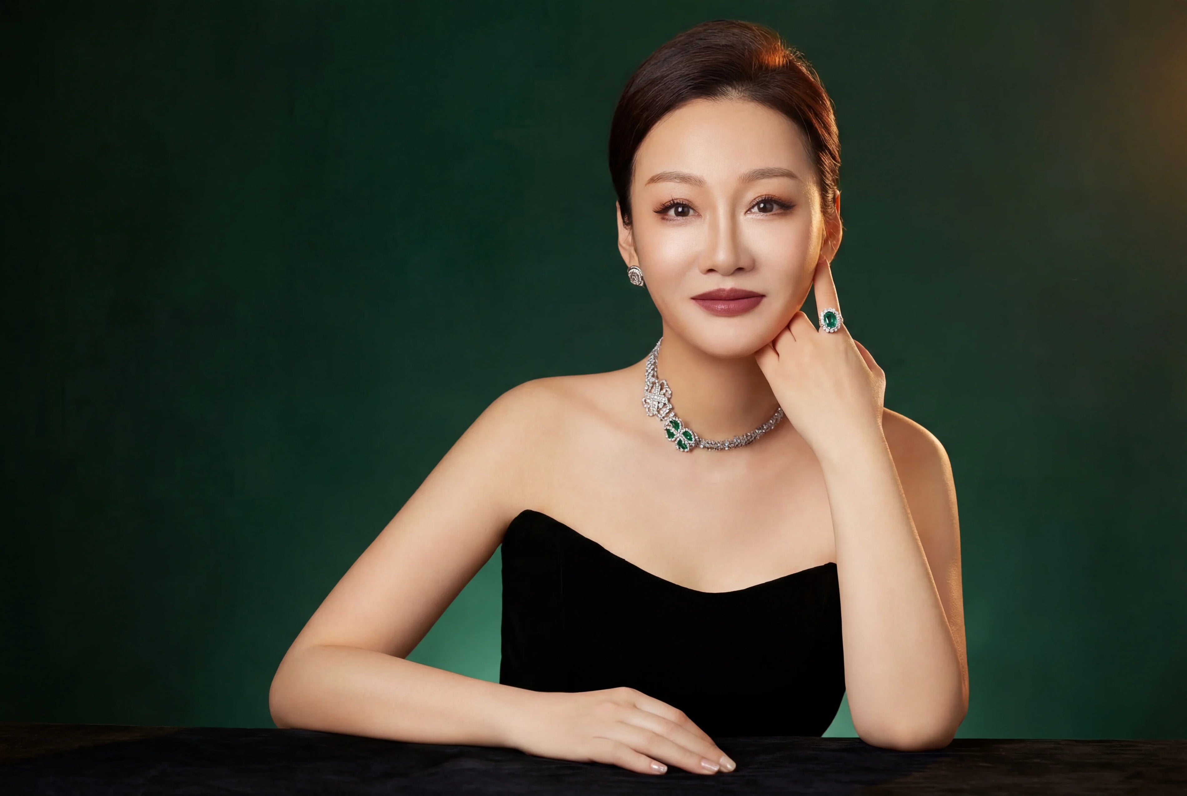 Woman in black strapless dress with elegant gemstone jewelry and natural manicure, seated against green background