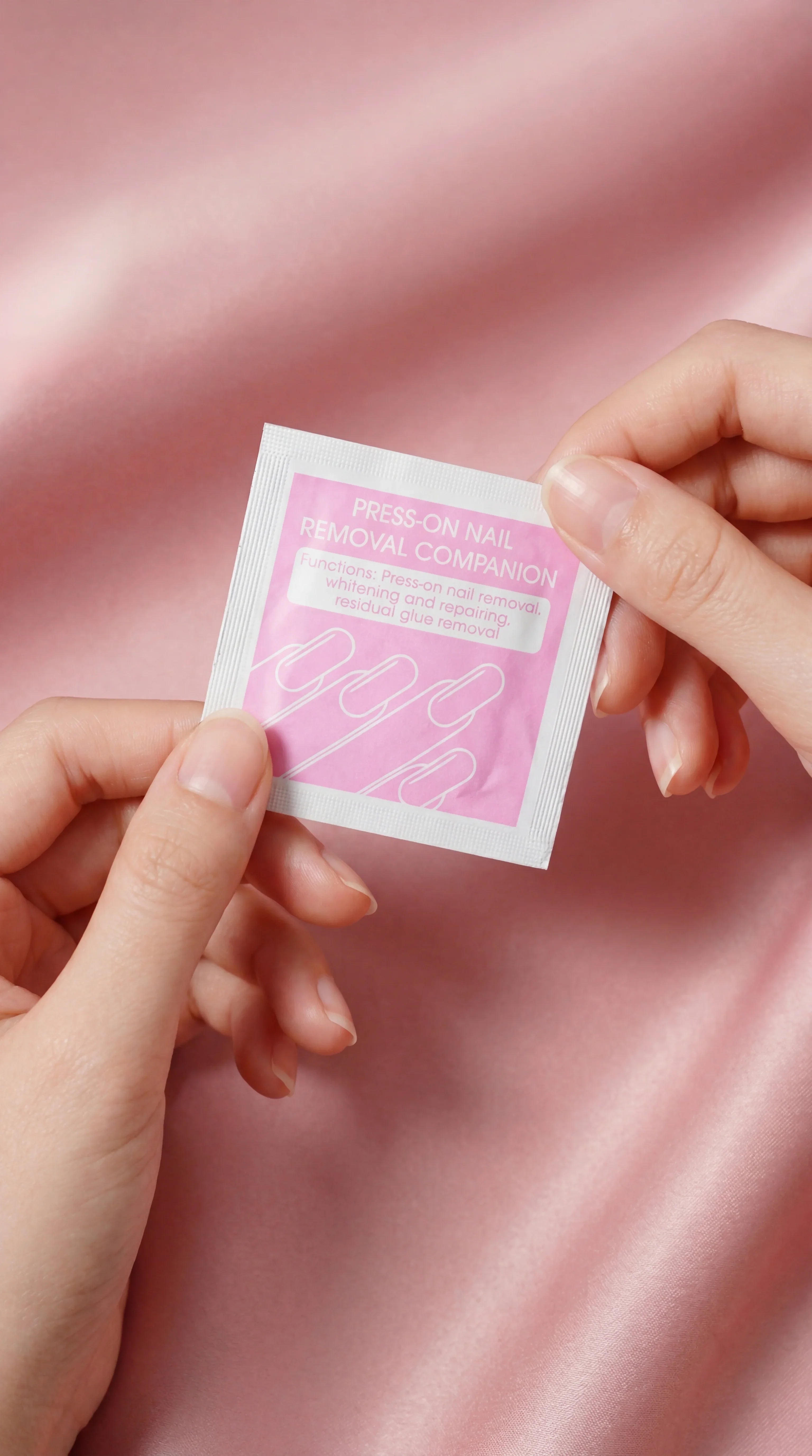 Hands holding a pink press-on nail removal wipe packet on a soft pink satin background.