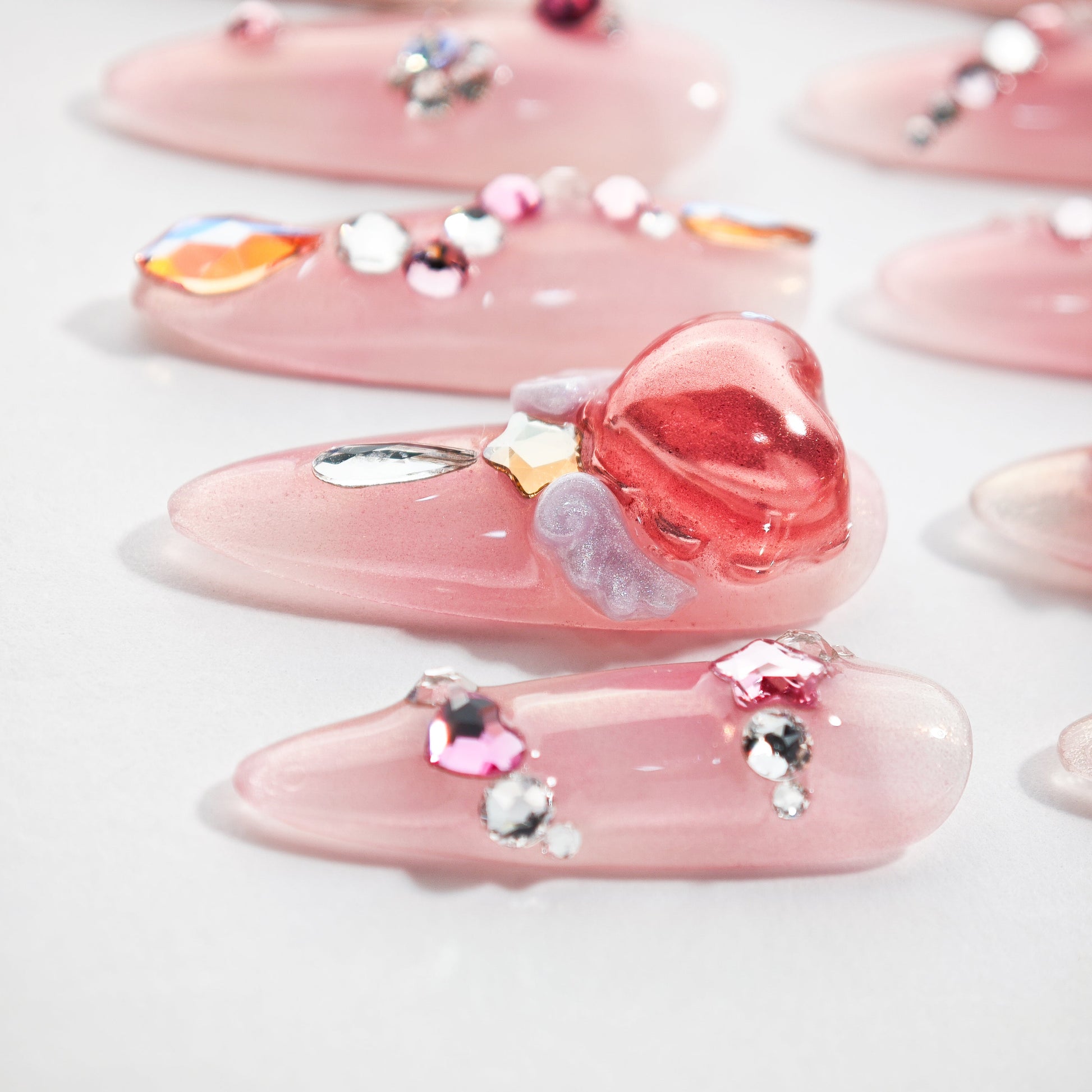 Pink press-on nails with 3D heart, rhinestones, and iridescent gem accents on white background