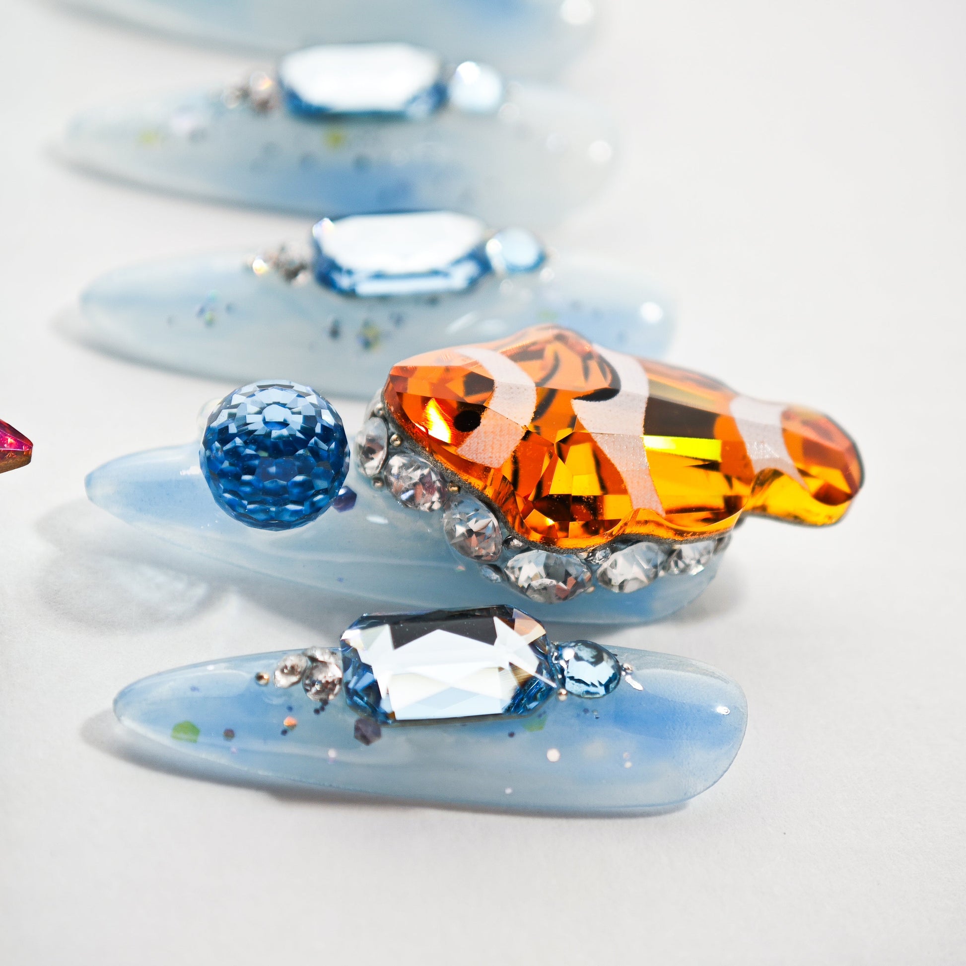 Blue press-on nails with large orange, blue, and clear gemstone accents and glitter details
