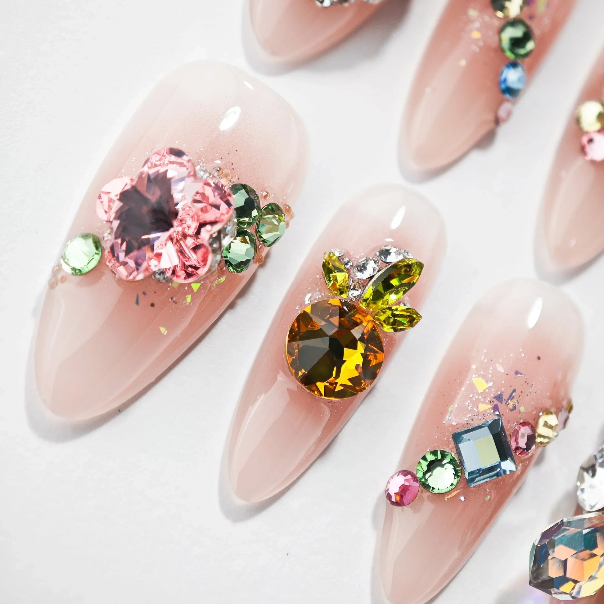 Long nude press-on nails with colorful gem and crystal embellishments, ombre design, glitter accents.