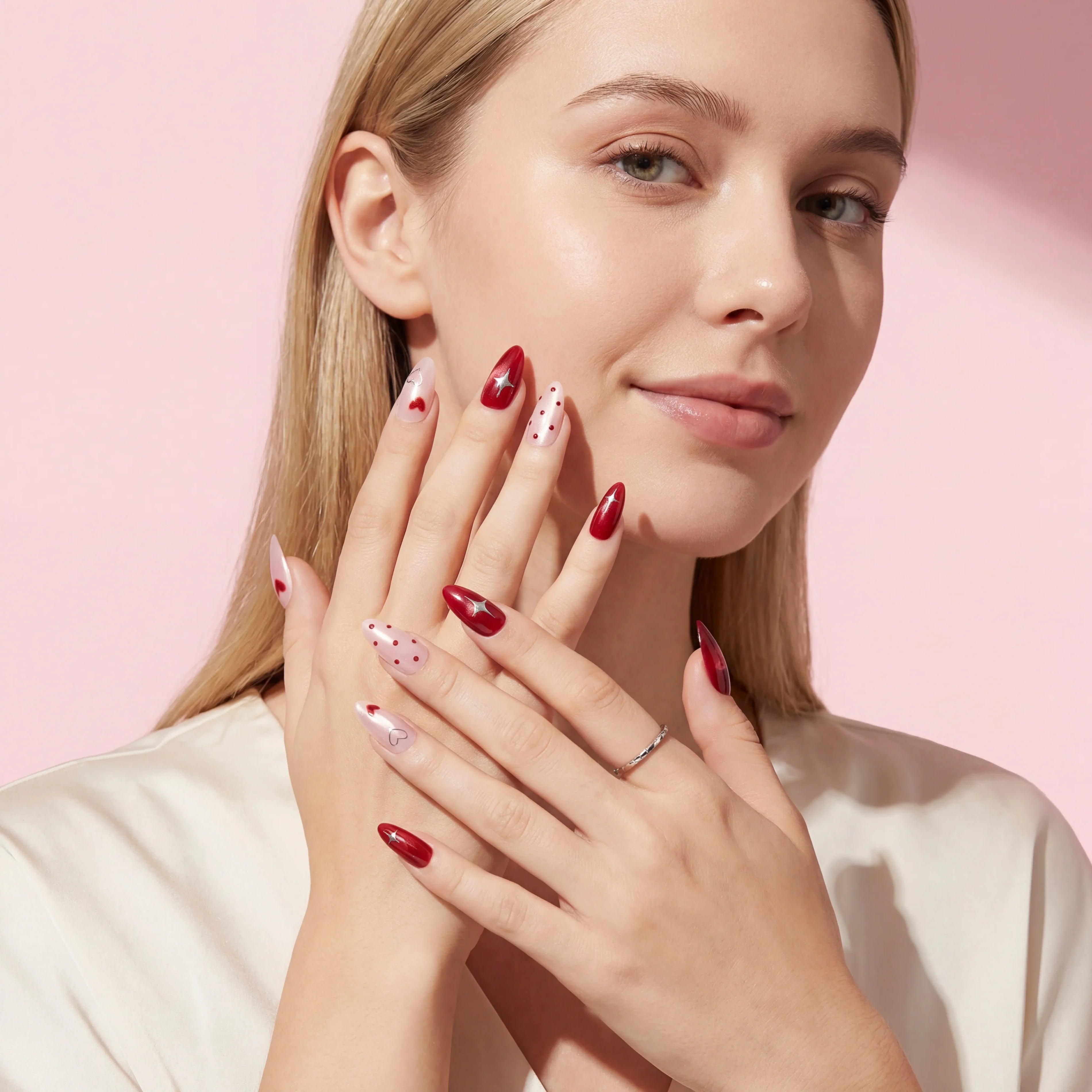 Red and white press-on nails with hearts, stars, and polka dots on woman's hands against pink background